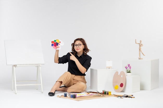 Front view young female sitting with paints and easel on a white background