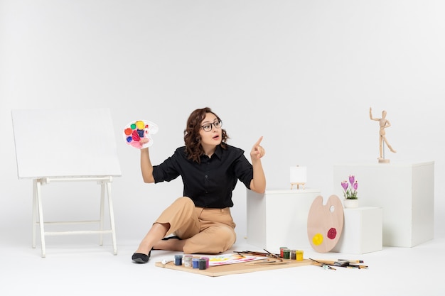 Free photo front view young female sitting with paints and easel on the white background