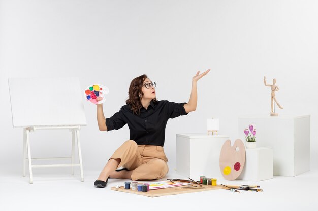 Front view young female sitting with paints and easel on white background