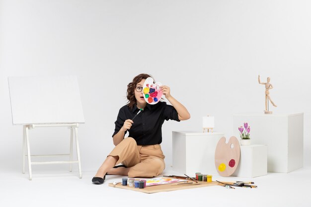 Front view young female sitting with paints and easel on white background