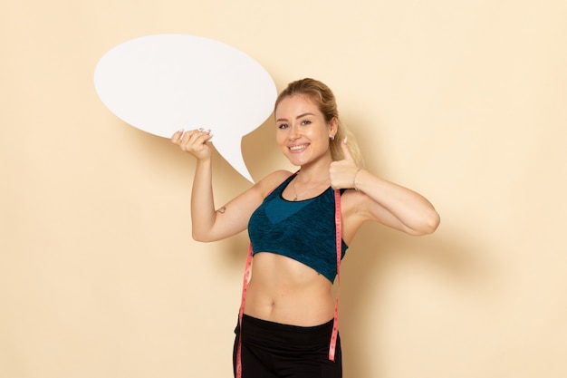 Free photo front view young female in sport outfit holding white sign and measuring her body on a white wall body sport beauty health exercises fit