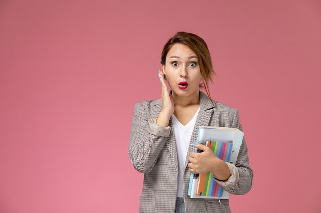 Front view young female student in grey coat with copybooks and shocked expression on pink background lessons university college study