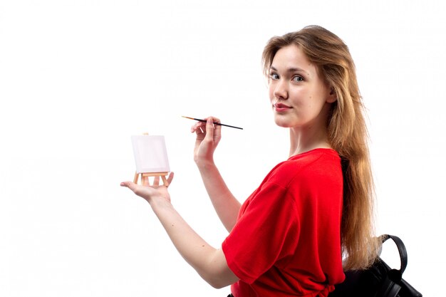 A front view young female student in red shirt black bag holding paintbrush painting smiling on the white