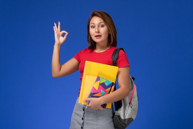 Front view young female student in red shirt wearing backpack holding files and copybook on blue background.