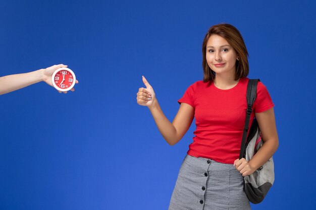 Front view young female student in red shirt wearing backpack showing like sign on light blue background.