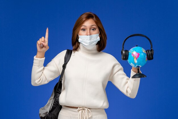 Front view young female student in white jersey wearing mask and backpack holding globe with headphones on the blue wall