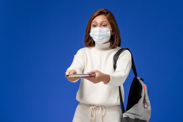 Front view young female student in white jersey wearing mask with bag and copybook thinking on blue wall