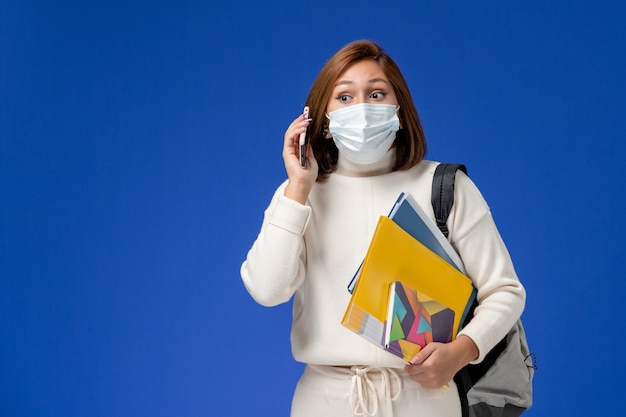 Front view young female student in white jersey wearing mask with bag and copybooks talking on the phone on blue wall