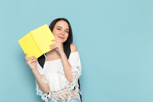 Front view young female student in white shirt blue jeans and black bag holding yellow copybook on the blue space female student university