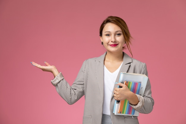 Front view young female student in white t-shirt grey coat and grey trousers with copybook in her hands smiling on the pink desk lessons university college study