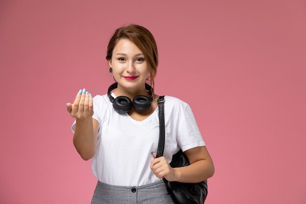 Front view young female student in white t-shirt and grey trousers with earphones and smiling expression on the pink background student lessons university college