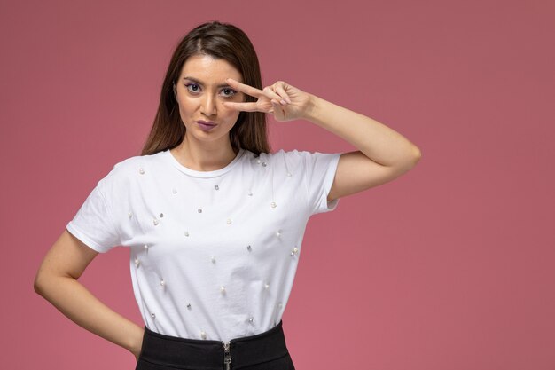 Front view young female in white shirt posing and showing victory sign on light-pink wall, woman model woman pose