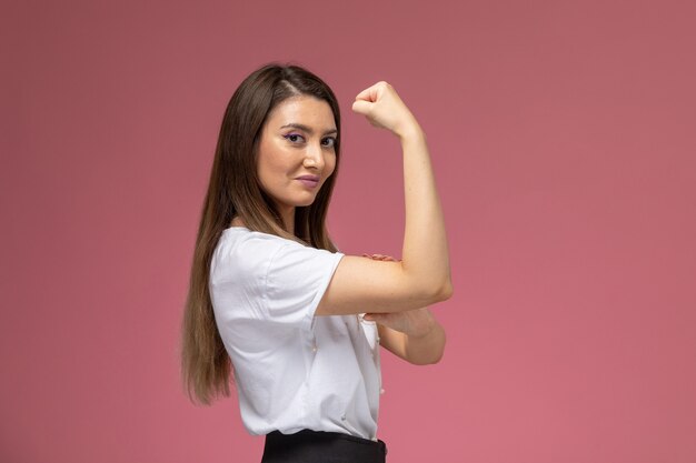 Front view young female in white shirt smiling and flexing on pink wall, model woman