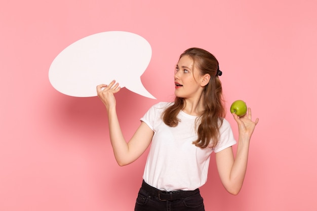 Front view of young female in white t-shirt holding fresh green apple and big white sign on light pink wall