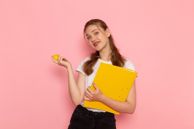 Front view of young female in white t-shirt holding fresh lemon with files smiling on the pink wall