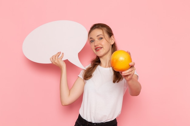 Free photo front view of young female in white t-shirt holding grapefruit and white sign smiling on pink wall