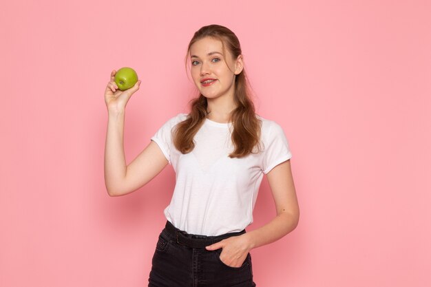 Front view of young female in white t-shirt holding green apple smiling on pink wall