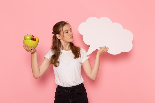 Free photo front view of young female in white t-shirt holding plate with fruits and white sign on the pink wall