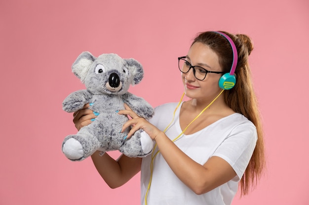 Free Photo front view young female in white t-shirt just listening to music via earphones and holding cute toy on the pink background 