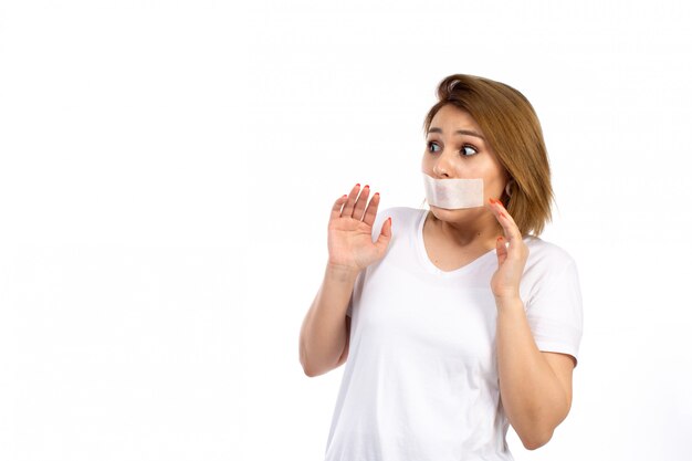 A front view young female in white t-shirt with white bandage around her mouth afraid of threats on the white