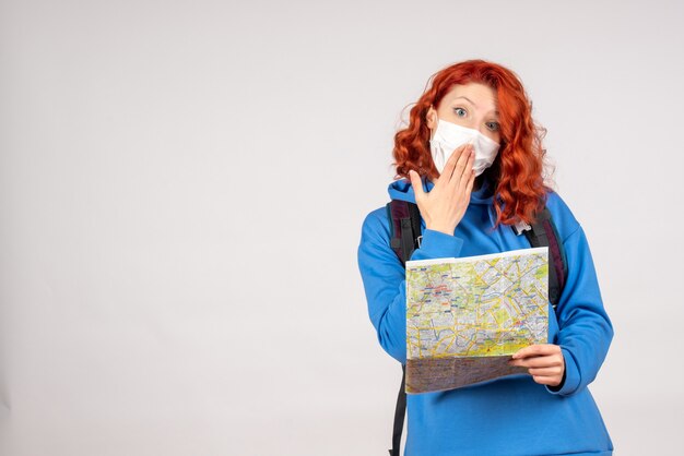 Front view of young female with backpack and map in mask on white wall