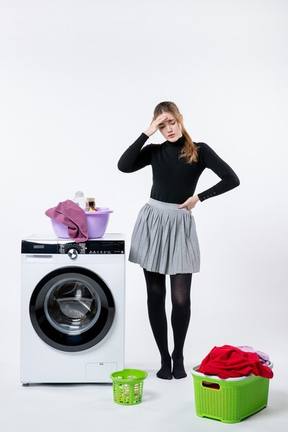 Front view of young female with washer and dirty clothes having headache on white wall