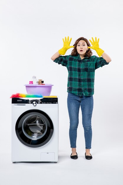 Front view of young female with washing machine in yellow gloves on white wall