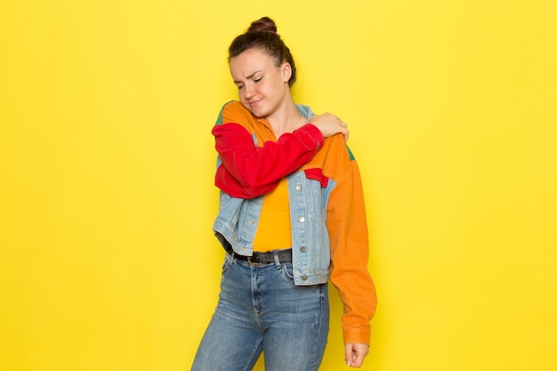 A front view young female in yellow shirt colorful jacket and blue jeans having a back ache