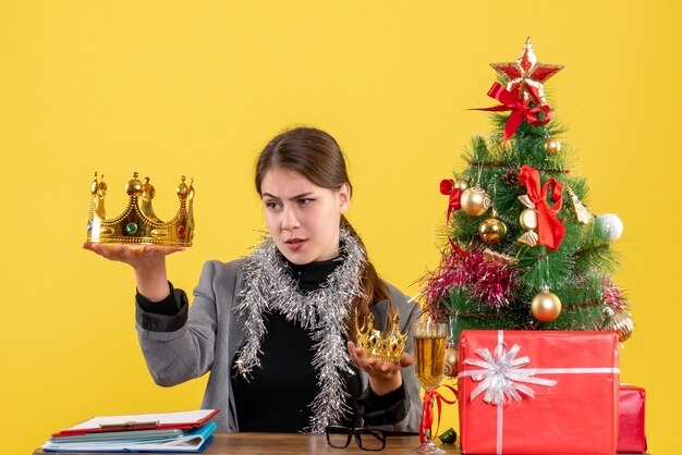 Front view young girl holding big and small crown and checking big one xmas tree and gifts cocktail