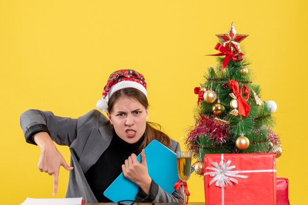 Front view young girl with xmas hat sitting at the table acting with anger xmas tree and gifts cocktail