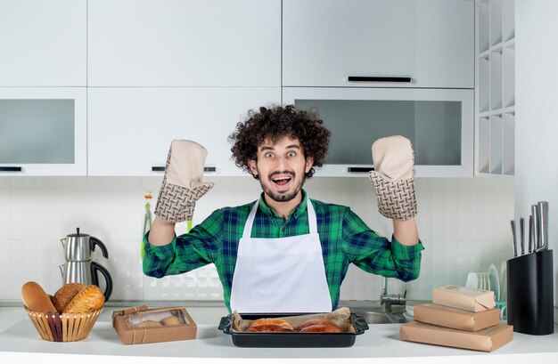 Front view of young guy wearing holder standing behind table freshly-baked pastry on it in the kitchen