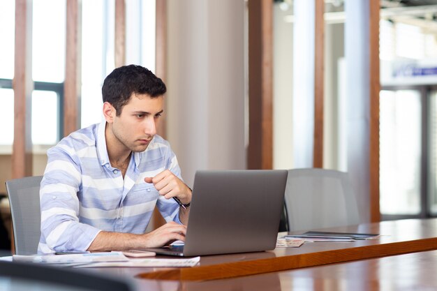 A front view young handsome man in striped shirt working inside conference hall using his silver laptop during daytime work activity building