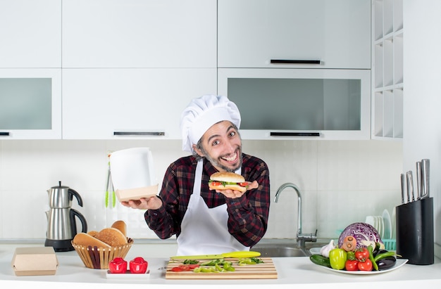 Front view of young hungry man holding up burger standing behind kitchen table