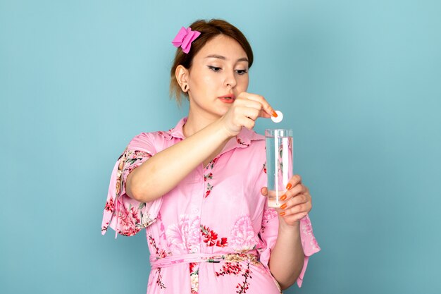 A front view young lady in flower designed pink dress putting pill into the glass of water on blue