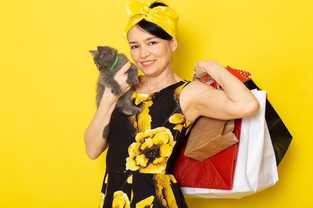 Free photo a front view young lady in yellow-black flower designed dress with yellow bandage on head holding shopping packages and kitten on the yellow