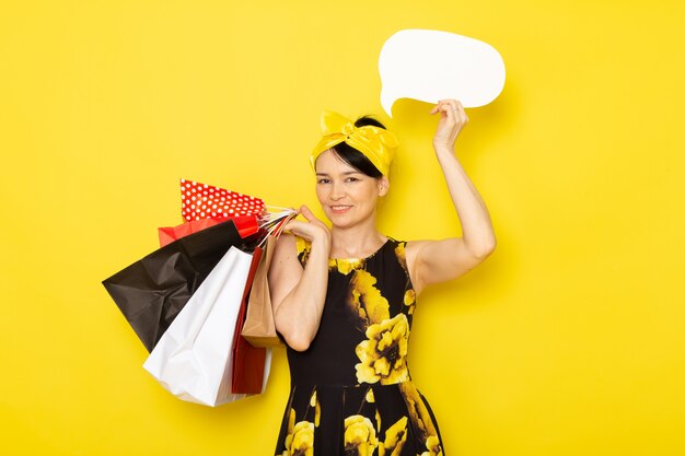 A front view young lady in yellow-black flower designed dress with yellow bandage on head holding shopping packages white sign on the yellow