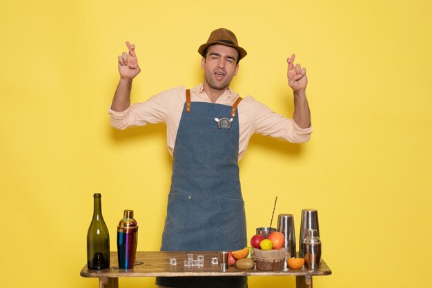 Front view young male bartender in front of table with drinks crossing his fingers on yellow background