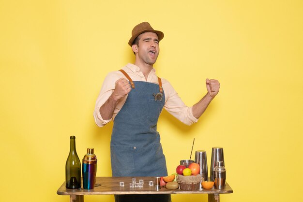 Front view young male bartender in front of table with shakers and drinks rejoicing on yellow background