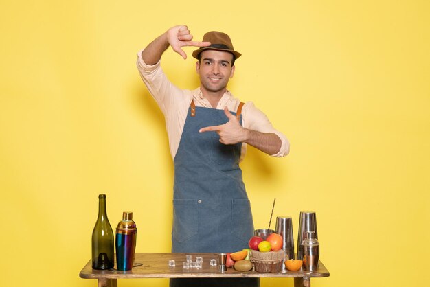 Front view young male bartender in front of table with shakers and drinks on a yellow background