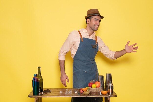 Front view young male bartender in front of table with shakers and drinks on the yellow background