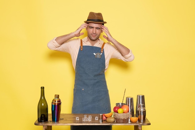 Front view young male bartender in front of table with shakers and drinks on yellow background