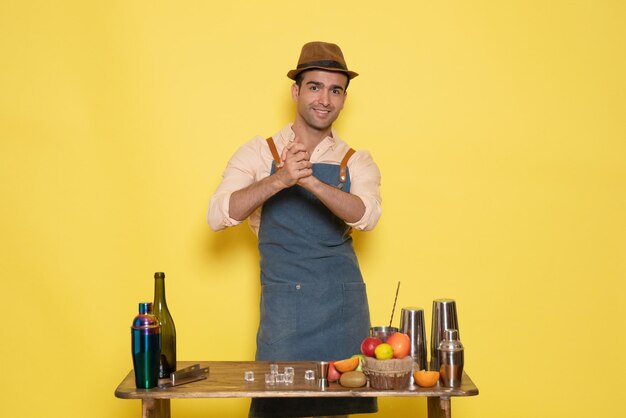 Front view young male bartender in front of table with shakers and drinks on yellow background