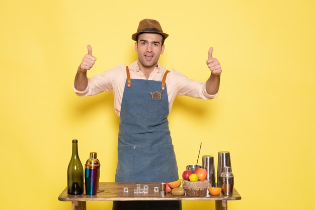 Front view young male bartender in front of table with shakers drinks on a yellow background