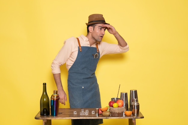 Free photo front view young male bartender in front of table with shakers drinks on yellow background