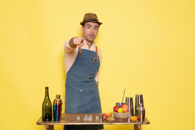 Free photo front view young male bartender in front of table with shakers drinks on yellow background