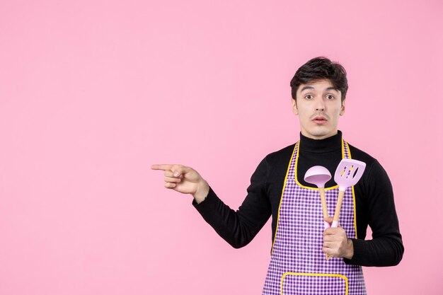 Front view young male in cape holding pink spoons on pink background profession food horizontal work cooking meal dough uniform color