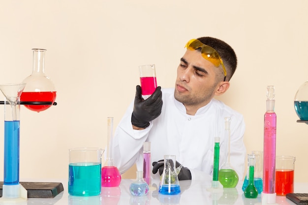 Free Photo front view young male chemist in white special suit sitting in front of table with solutions holding flask and thinking on cream desk lab chemistry science