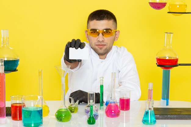 Front view young male chemist in white suit in front of table with colored solutions holding white card on the yellow wall science work lab chemistry