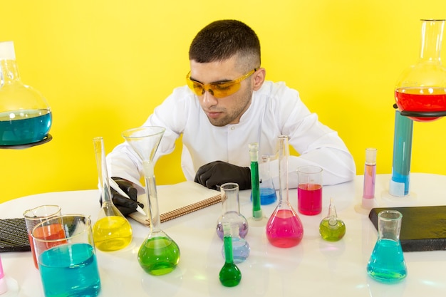 Front view young male chemist in white suit in front of table with colored solutions writing down notes on the yellow wall science work chemistry lab