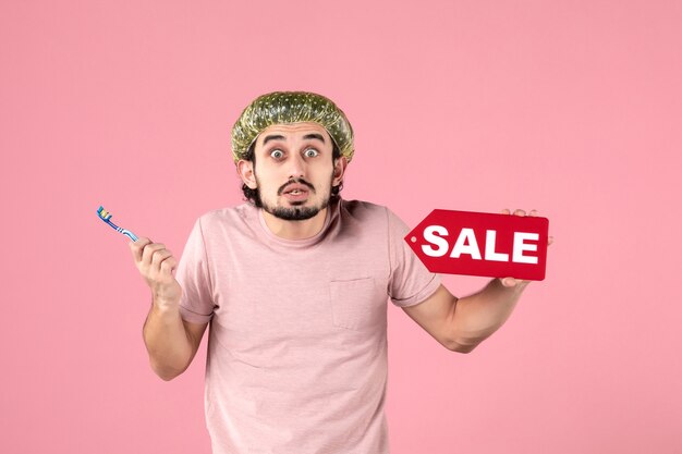 front view young male cleaning his teeth and holding sale nameplate on pink background